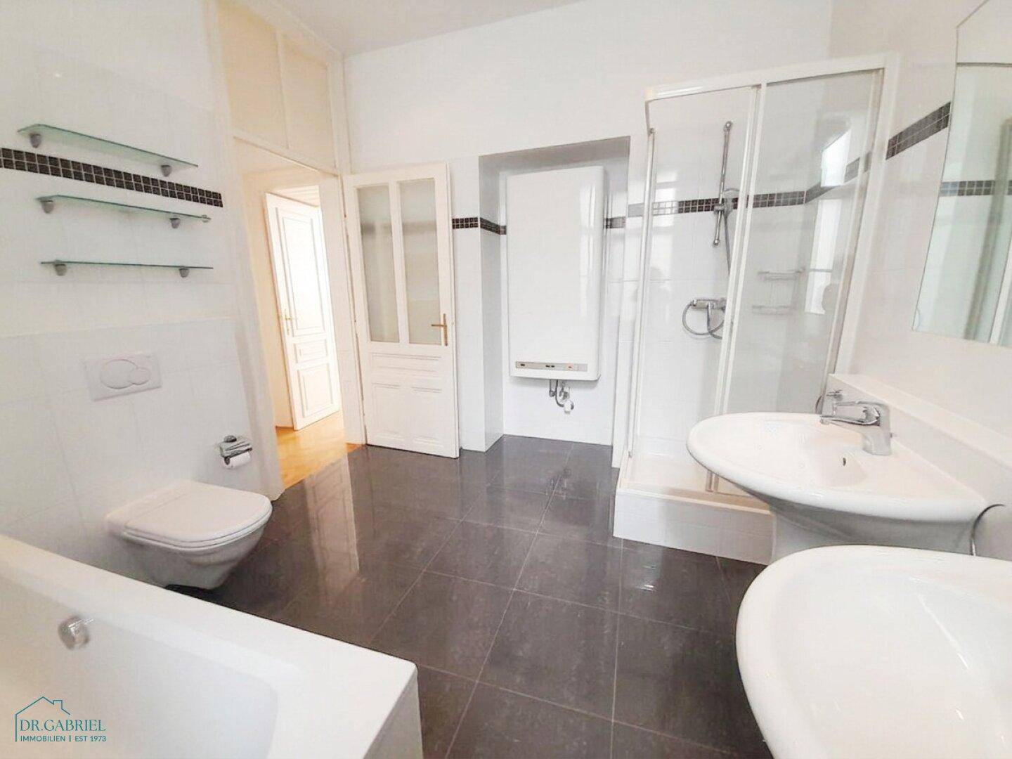 Badezimmer mit Badewanne, Dusche und Waschbecken, ausgestattet mit weißen Fliesen und dunklem Boden.
