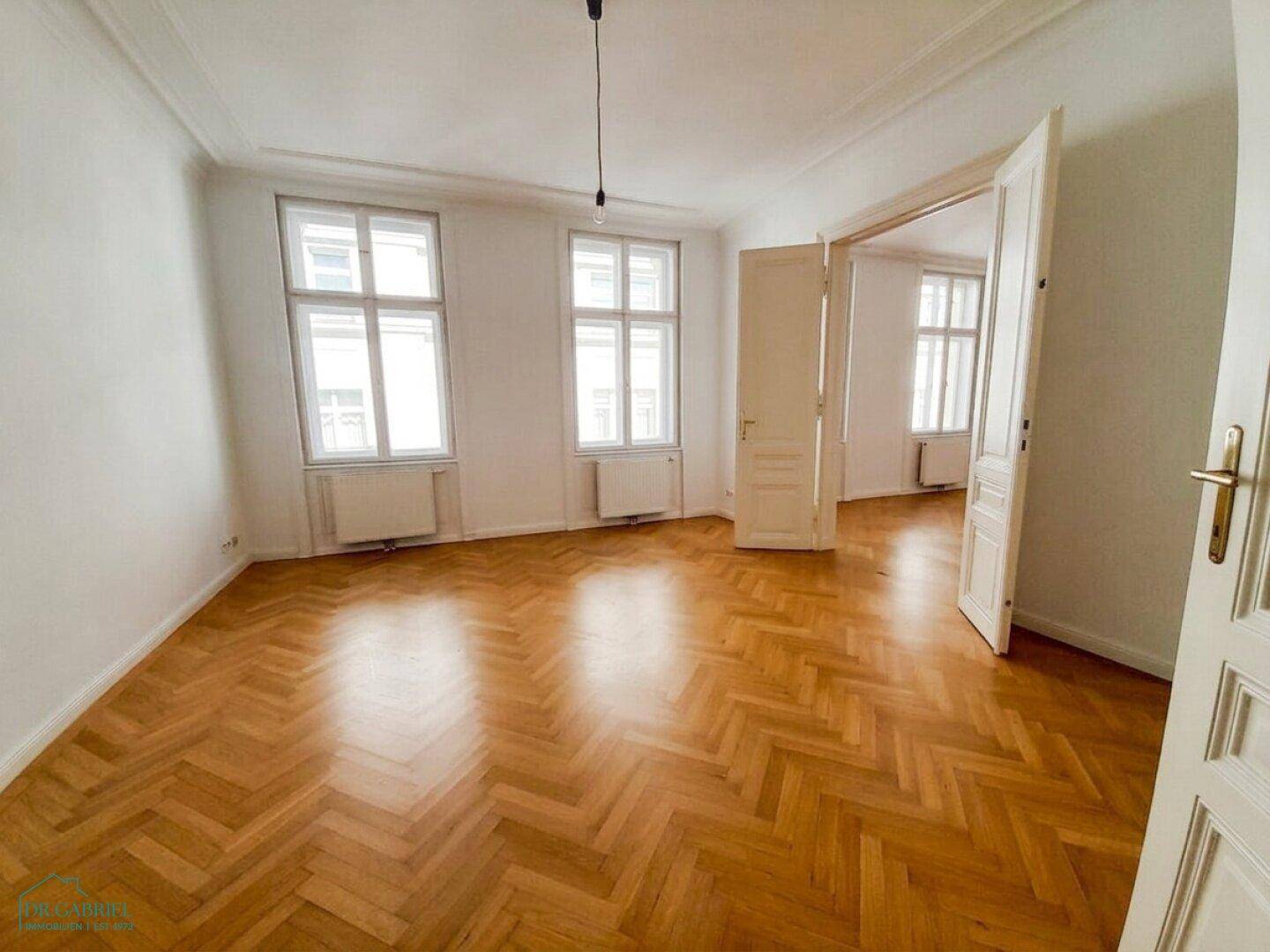 Geräumiges Zimmer mit Fischgrätparkett, hohen Decken und Zugang zu weiteren Räumen.