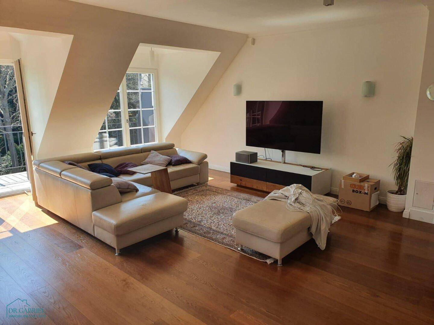 Großes Wohnzimmer mit Holzboden, beiger Ledercouch und Blick ins Grüne durch die Fenster.