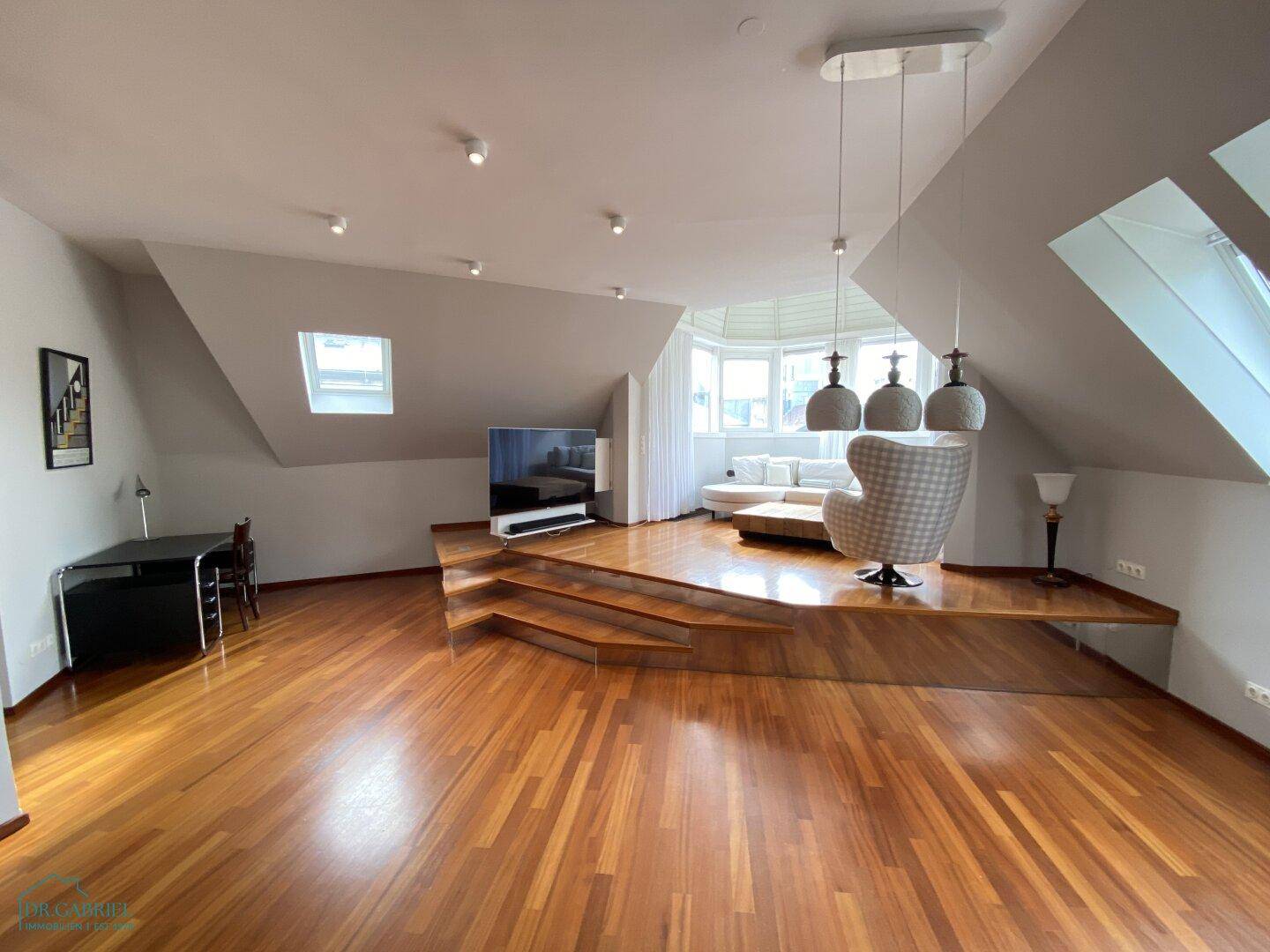 Weitläufiges Wohnzimmer mit Holzboden, Arbeitsbereich und gemütlicher Sitzecke unter Dachschrägen.