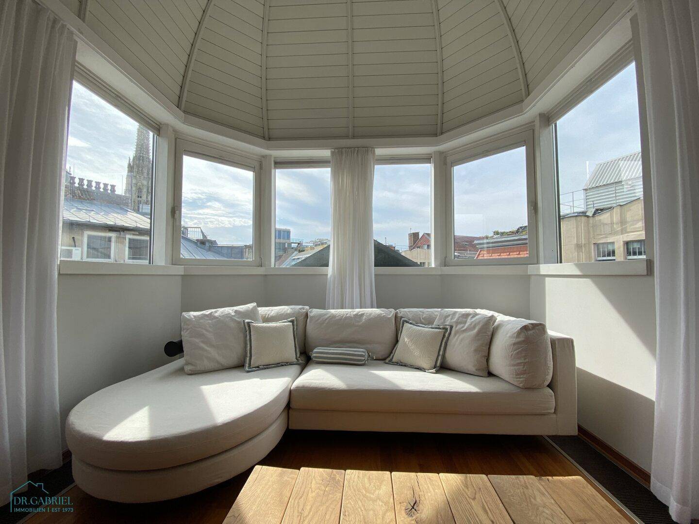 Helles Wohnzimmer mit gemütlichem Sofa in einem Erker mit vielen Fenstern und Stadtblick.