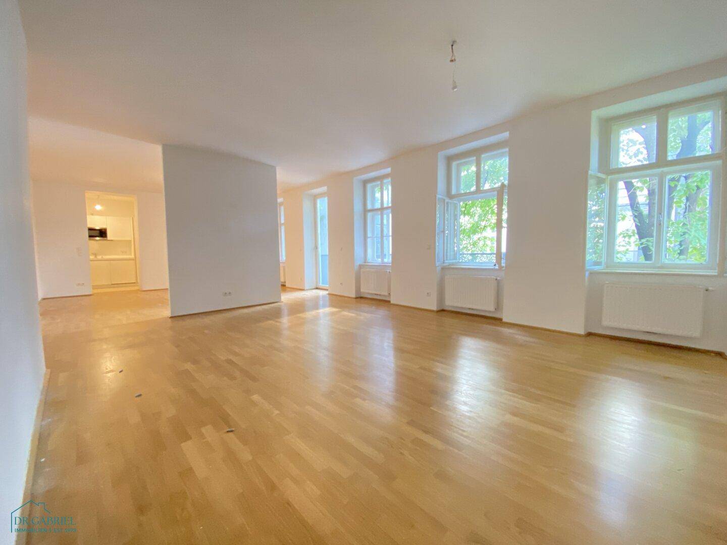 Helles, leeres Wohnzimmer mit glänzendem Parkettboden und hohen Fenstern, die viel Tageslicht hereinlassen.