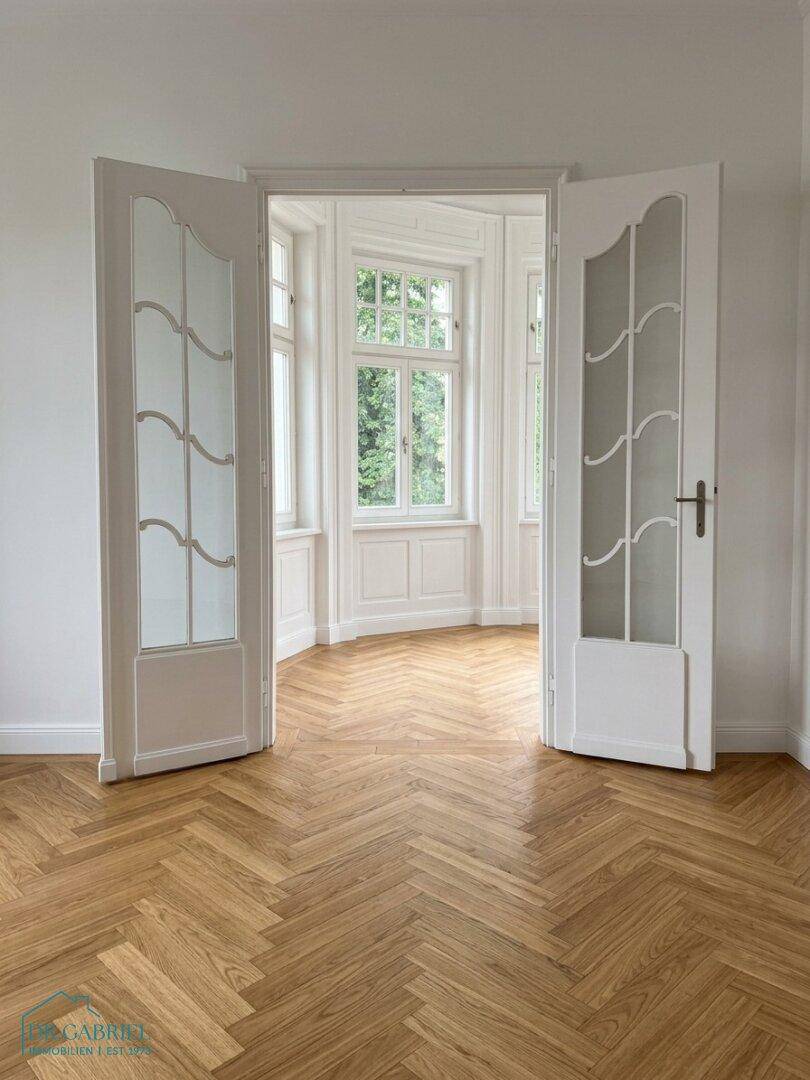 Heller Raum mit eleganten Flügeltüren, Fischgrätparkett und einem lichtdurchfluteten Erkerbereich.
