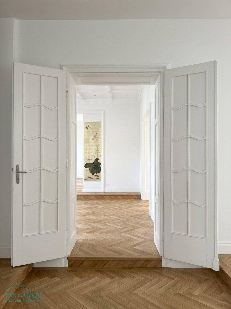 Elegante Flügeltüren öffnen sich zu einem weiteren Raum mit klassischem Fischgrätparkett und hohen Decken.
