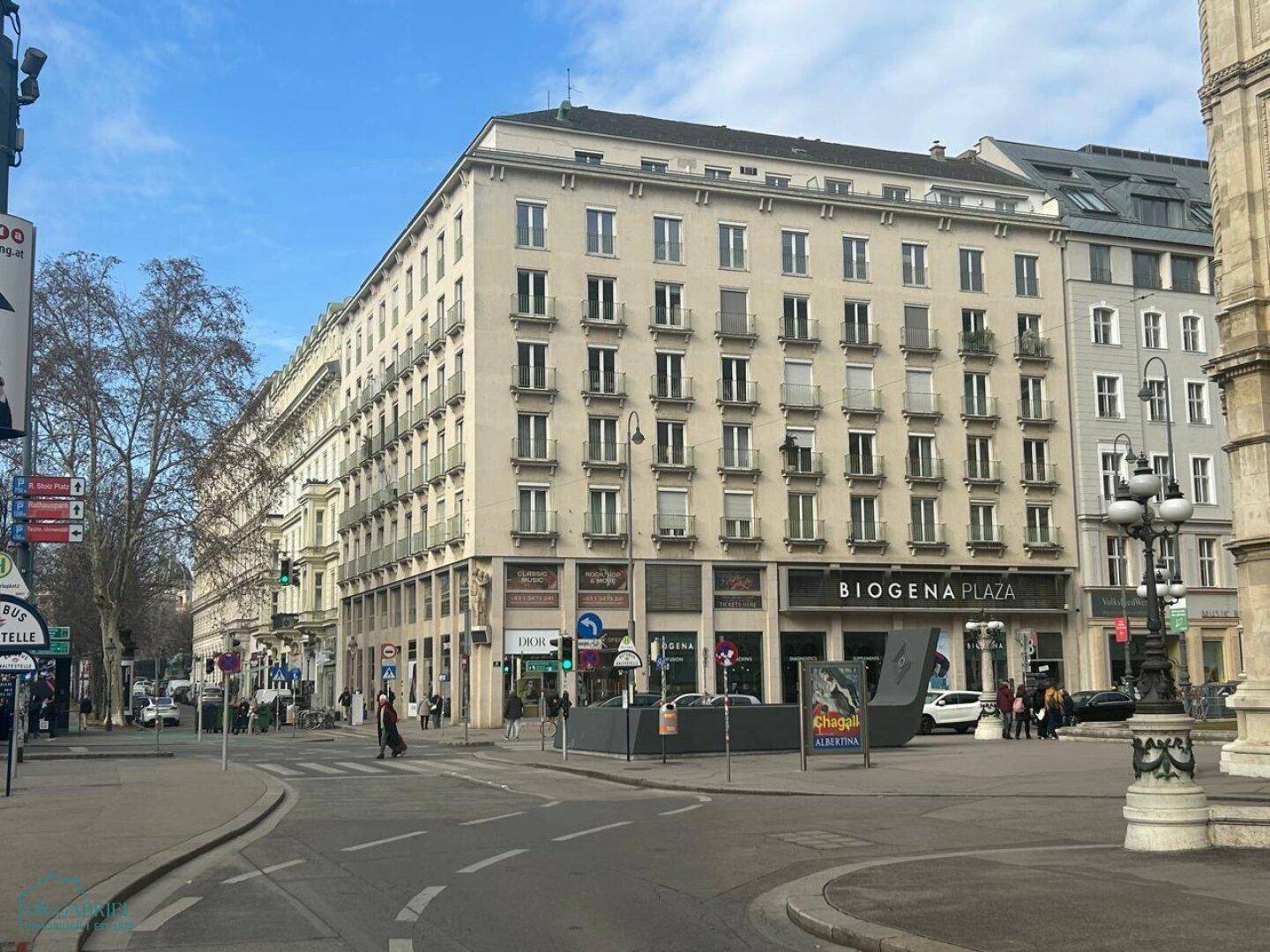 Großes Mehrfamilienhaus mit vielen Fenstern und kleinen Balkonen an einer belebten Stadtstraße.