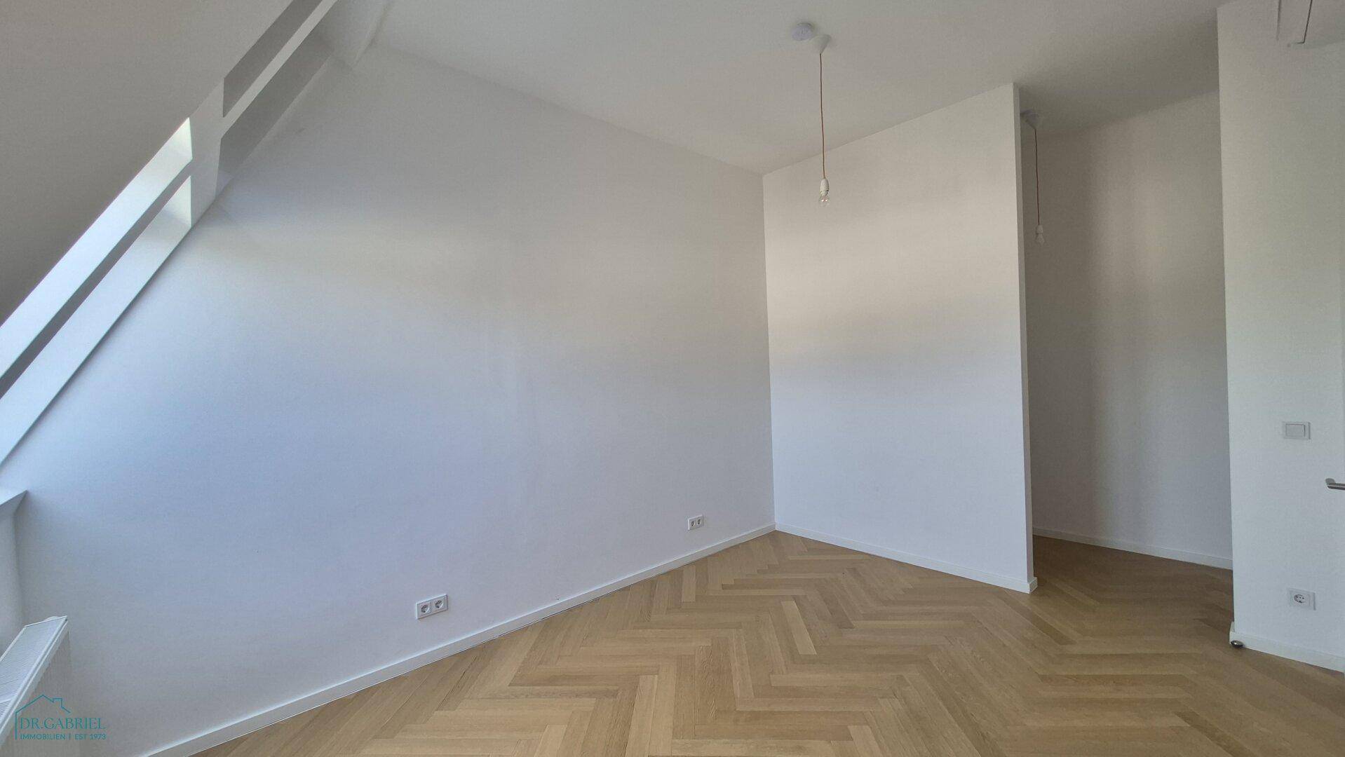 Geräumiger Raum mit Fischgrätparkett und schrägen Wänden, beleuchtet durch ein großes Dachfenster.