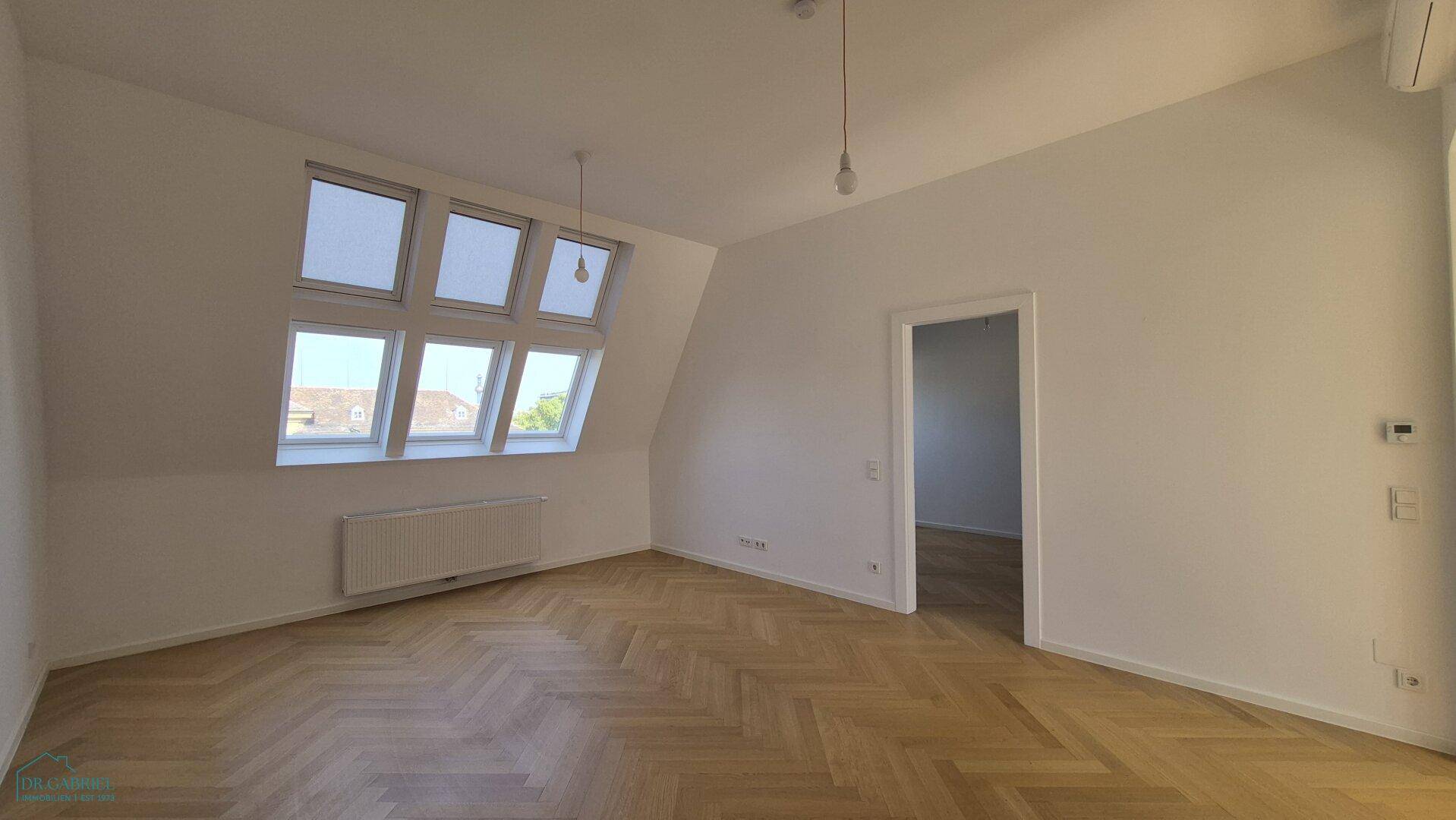 Heller, leerer Raum mit Fischgrätparkett, großen Dachfenstern und einem Heizkörper.
