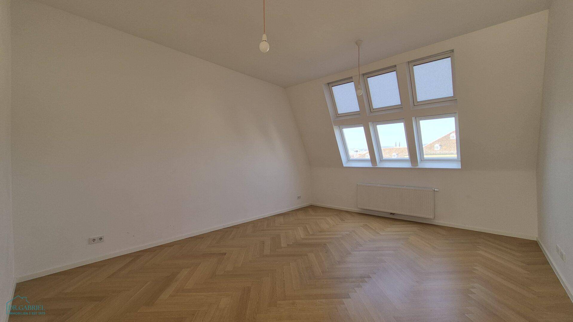 Geräumiger, unmöblierter Raum mit hellem Fischgrätparkett und vielen Dachfenstern für viel Tageslicht.
