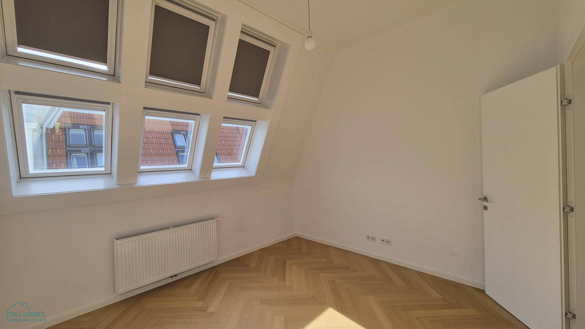 Heller, leerer Raum mit Fischgrätparkett, Dachfenstern und einem Heizkörper, ideal als Schlafzimmer.