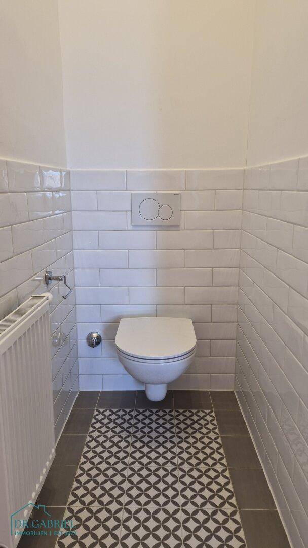 Modernes WC mit wandhängender Toilette, weißen Fliesen und gemustertem Fliesenboden.