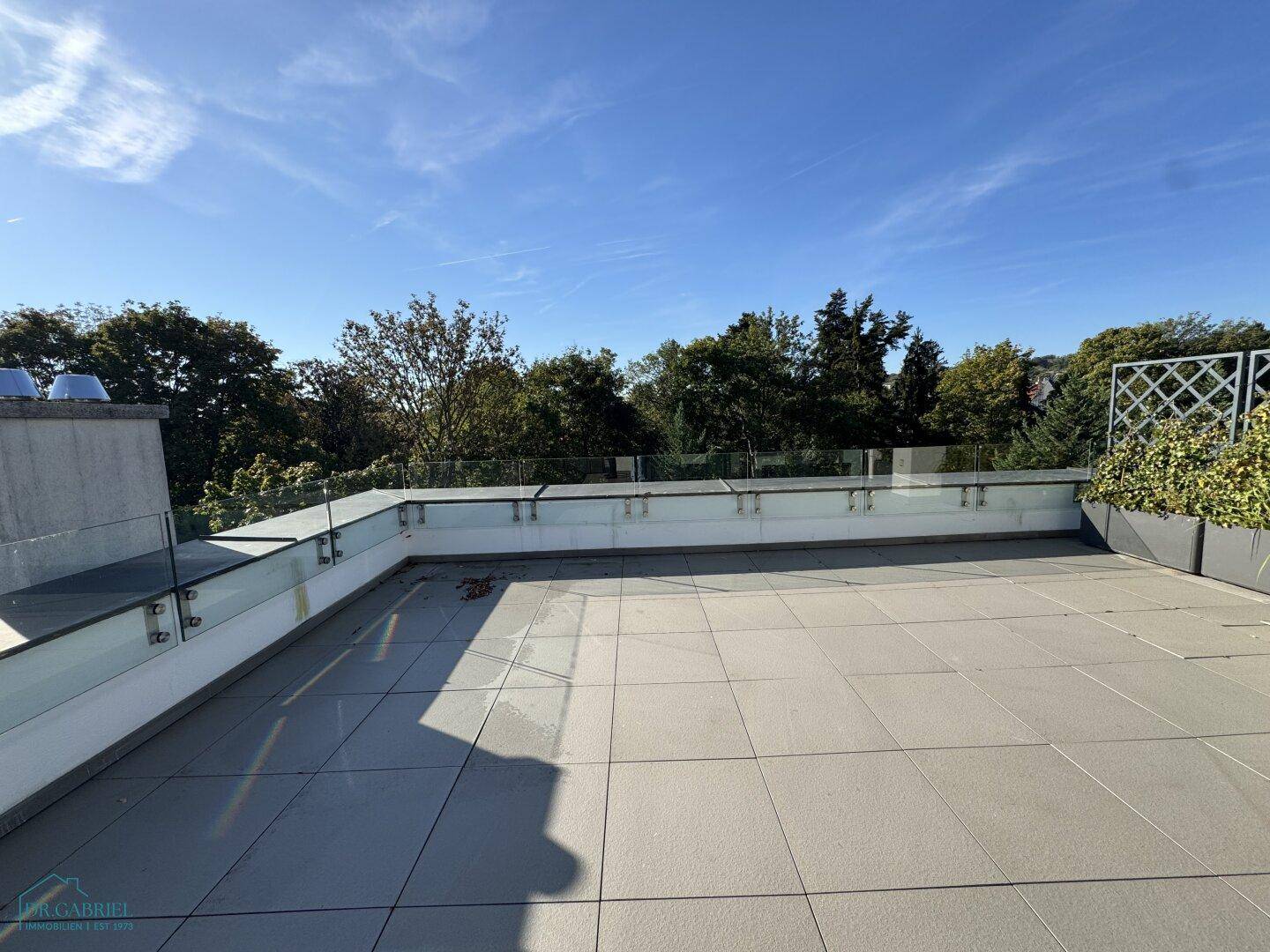 Sonnige Dachterrasse mit Fliesenboden und modernem Glasgeländer, umgeben von Bäumen.