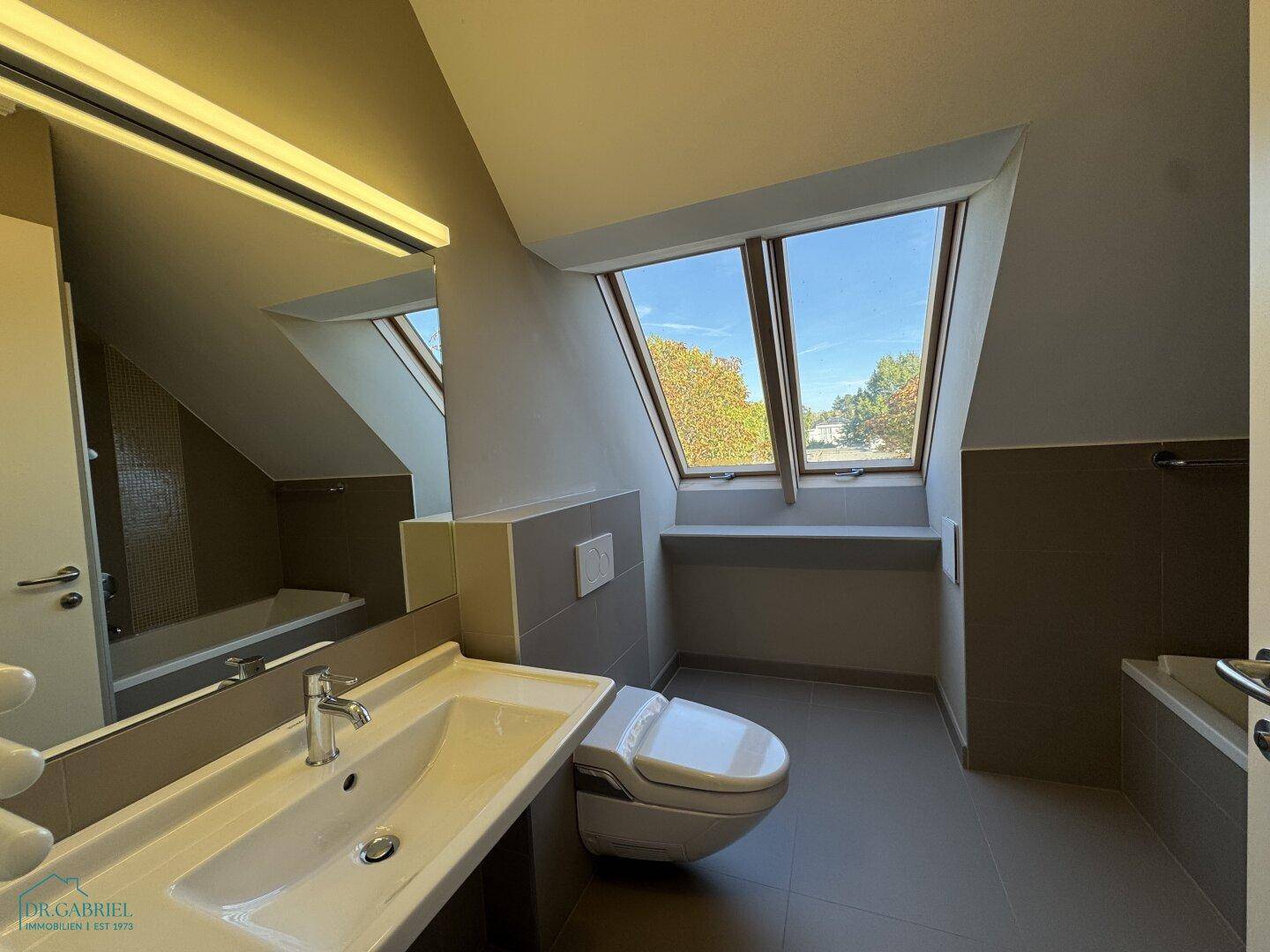 Helles Badezimmer mit Waschbecken, Toilette und Badewanne unter einem Dachfenster.