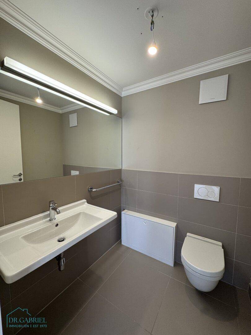 Helles Badezimmer mit Einzelwaschbecken, großem Spiegel und wandhängendem WC auf grauen Fliesen.