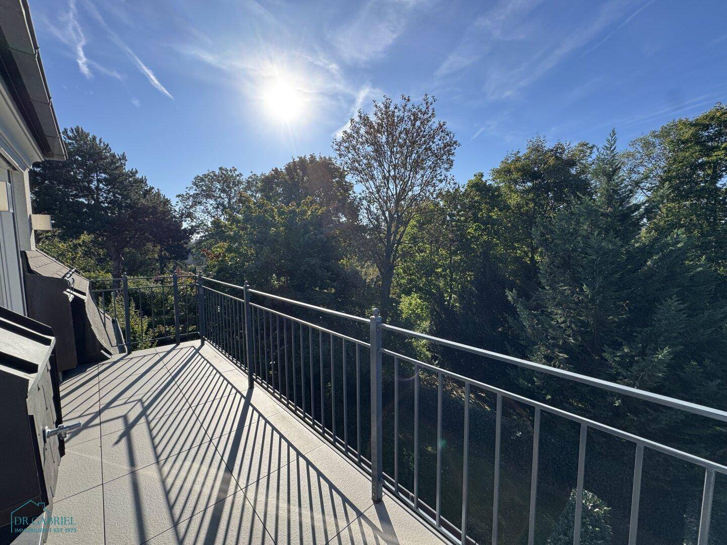 Heller Balkon mit Blick auf grüne Bäume und viel Sonnenschein.