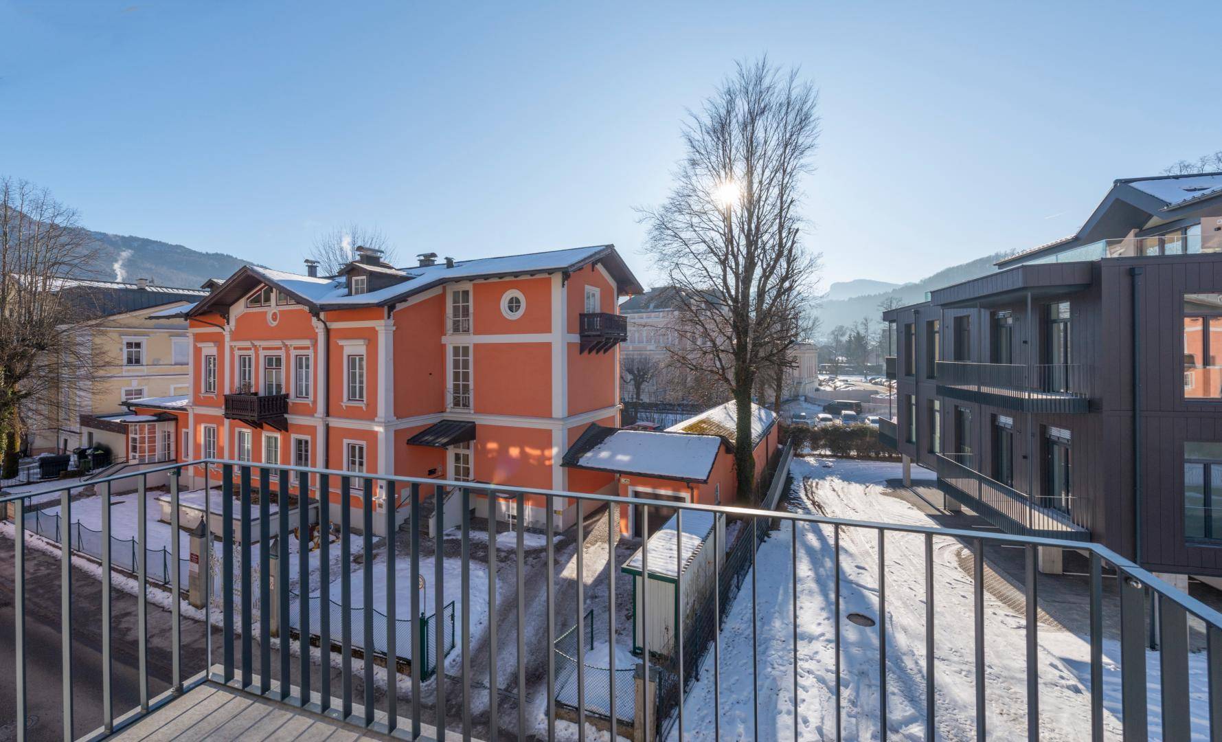 Winterlicher Ausblick vom Balkon auf eine verschneite Straße, Gebäude und Berge im Hintergrund.