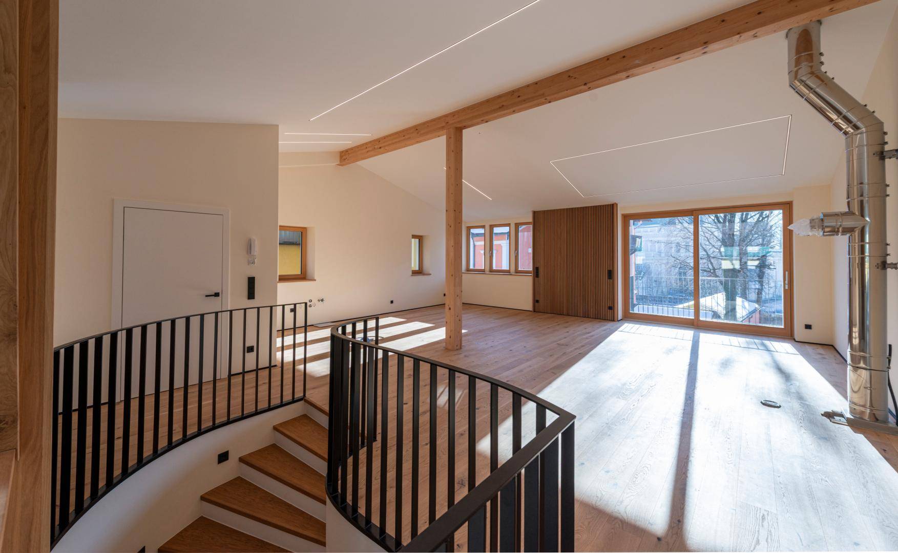 Heller, unmöblierter Wohnbereich mit Holzbalken, großer Schiebetür und moderner Treppe.