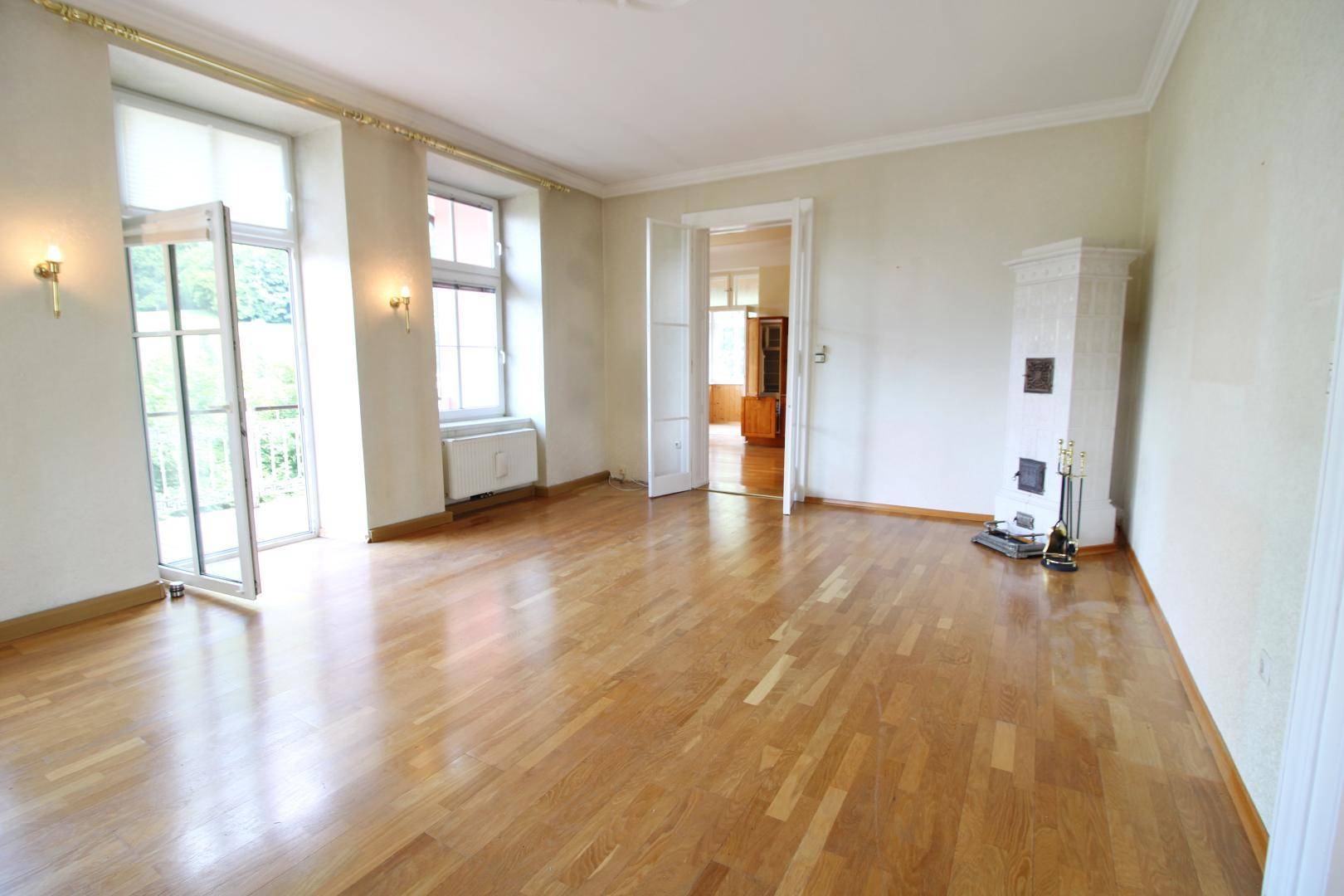 Großes, leeres Wohnzimmer mit Holzboden, Kachelofen und Zugang zu einem französischen Balkon.