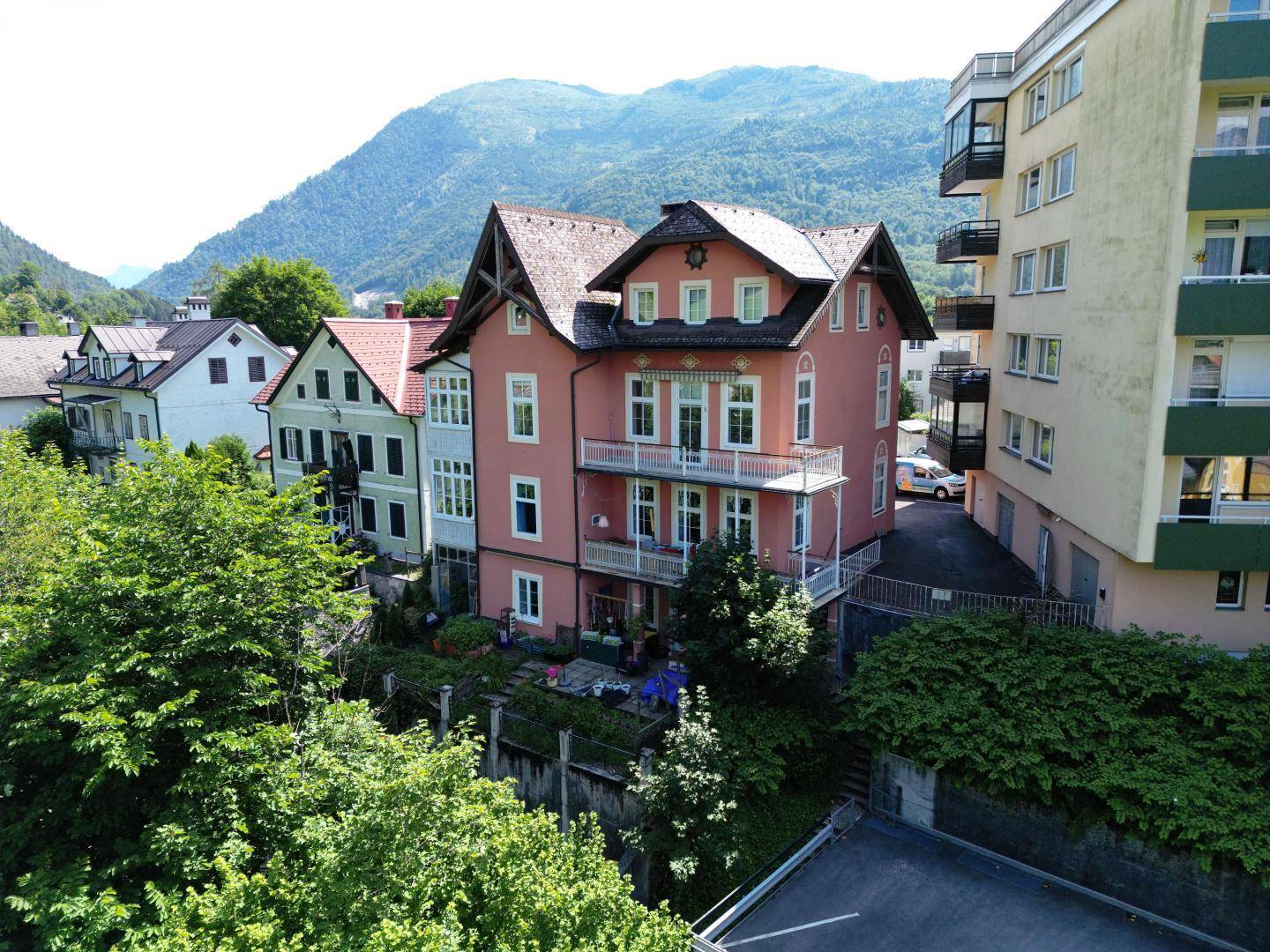 Historisches Gebäude mit rosafarbener Fassade, Balkonen und Bergblick in grüner Umgebung.