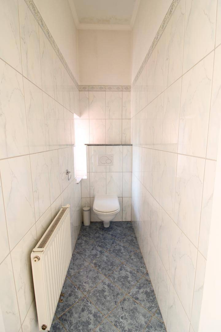 Schmale Gästetoilette mit weißen Fliesen, blaugrauem Boden und Wand-WC.