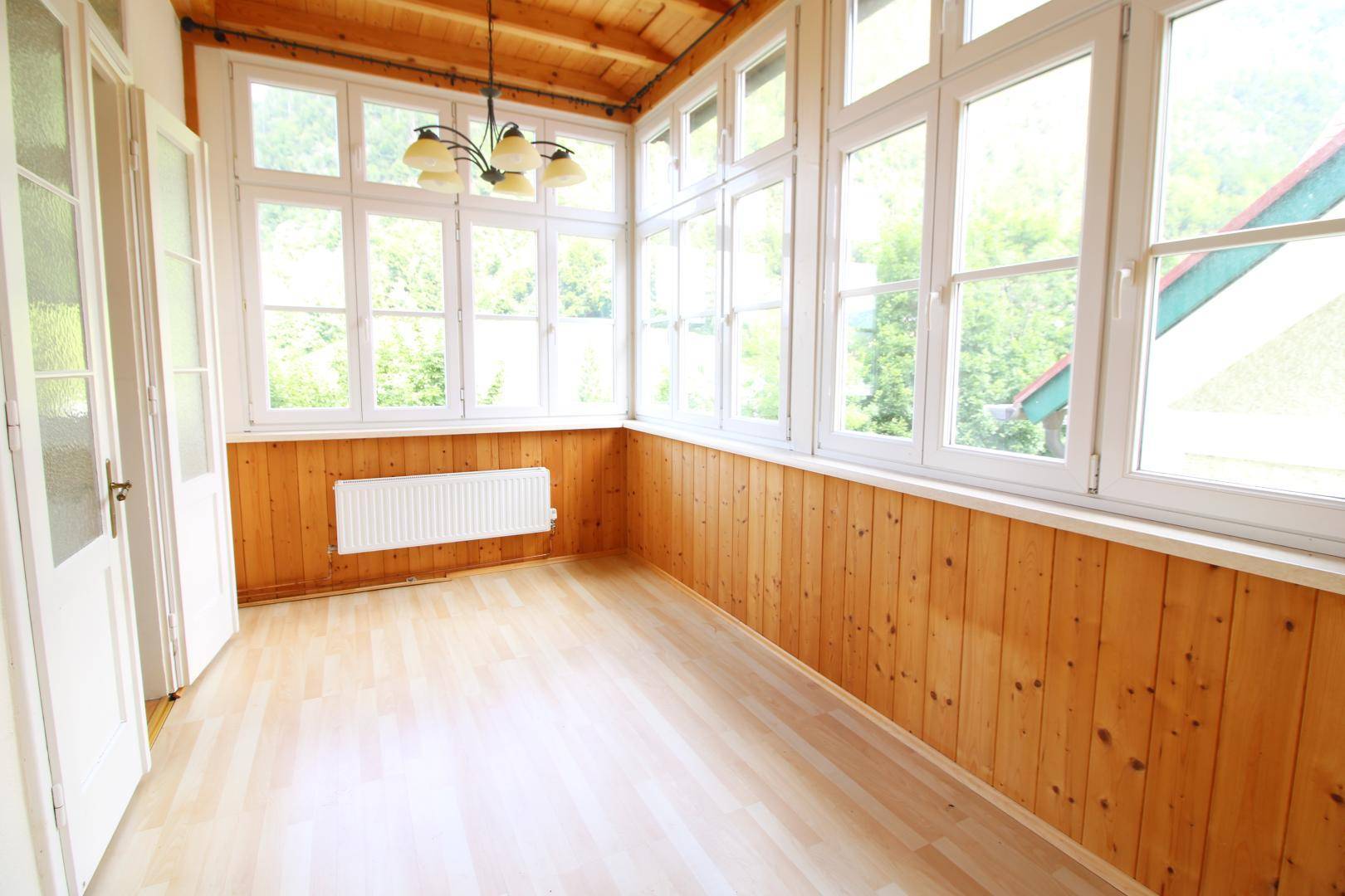 Heller Wintergarten mit Holzverkleidung, großen Fenstern und Blick ins Grüne.