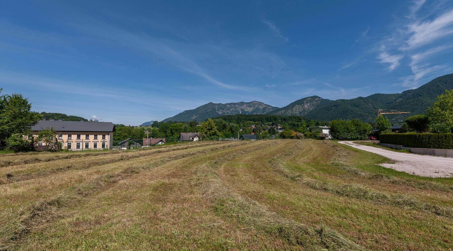Offenes Feld mit Heu und einer malerischen Bergkulisse im Hintergrund, umgeben von ländlicher Bebauung.