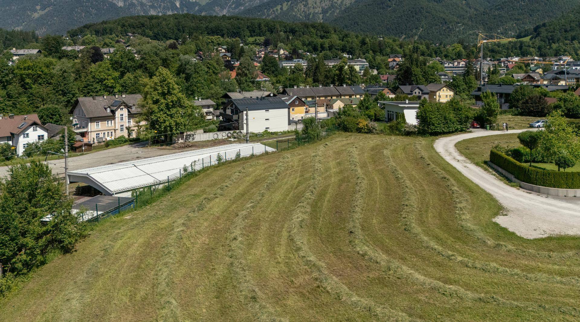 Weitläufiges Feld mit Heu und Blick auf eine Ortschaft mit Bergen im Hintergrund unter klarem Himmel.