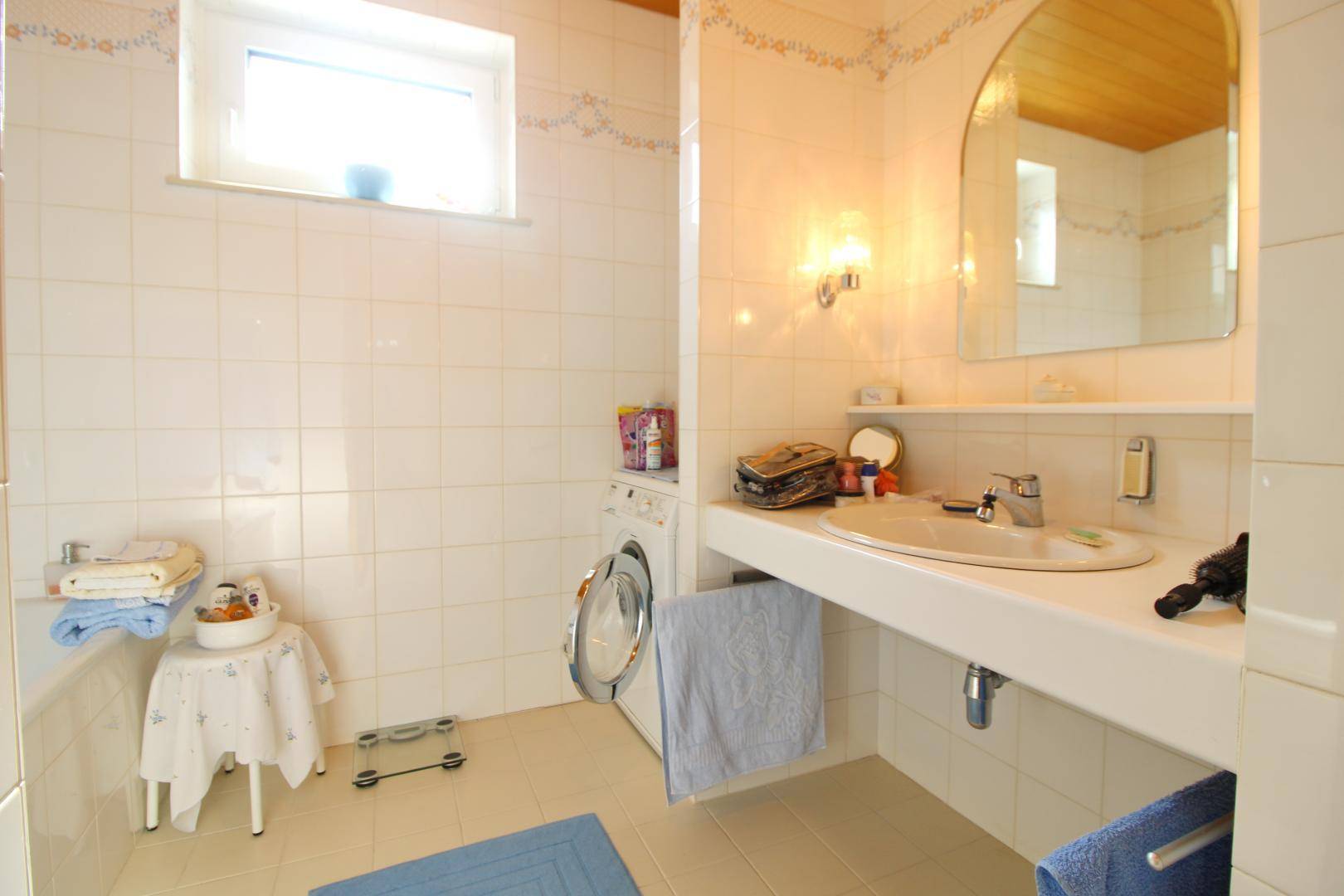 Funktionales Badezimmer mit weißen Fliesen, Waschbecken, Waschmaschine und Fenster.