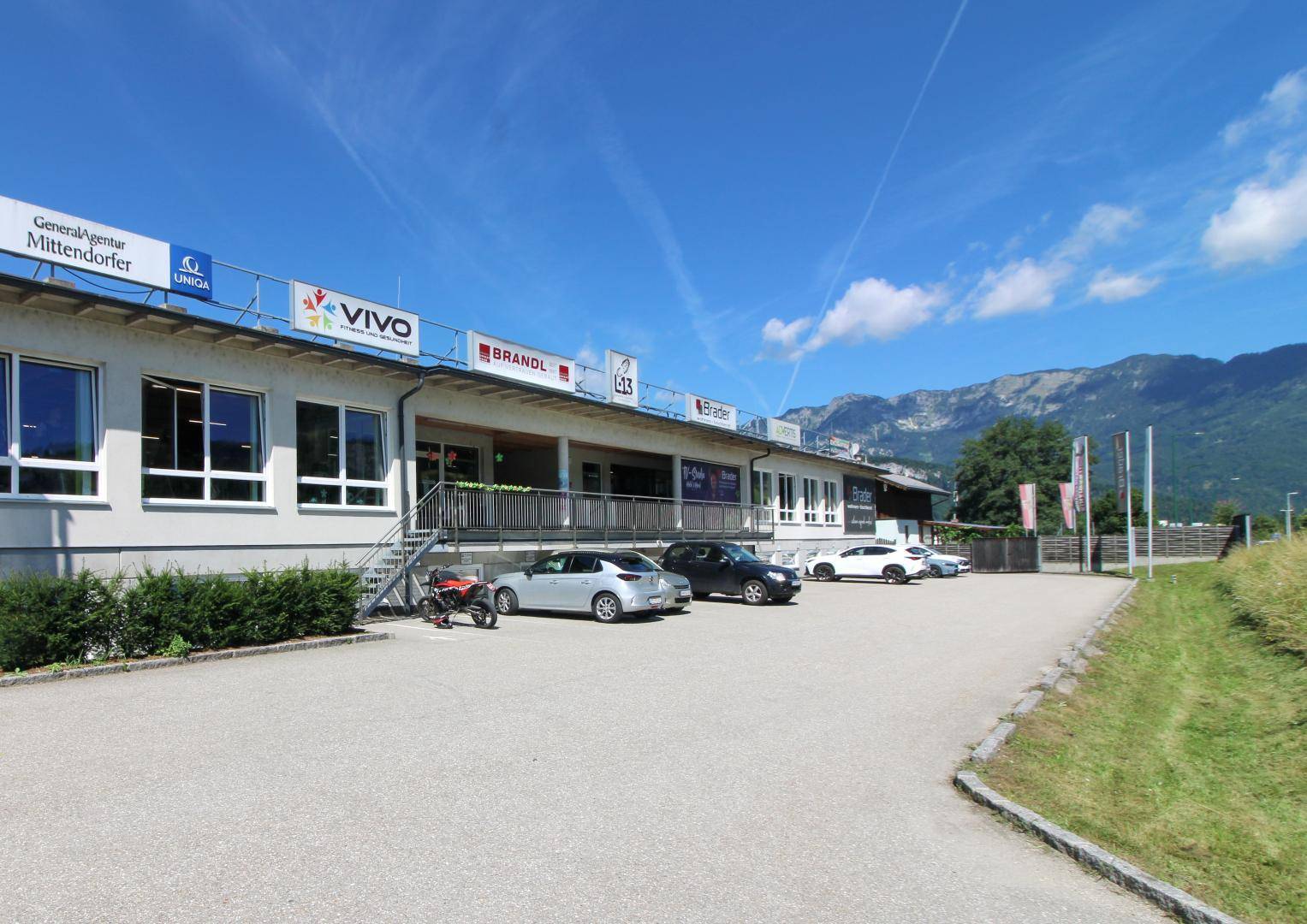 Fassade eines Geschäftsgebäudes mit Parkplätzen und Bergen im Hintergrund unter blauem Himmel.