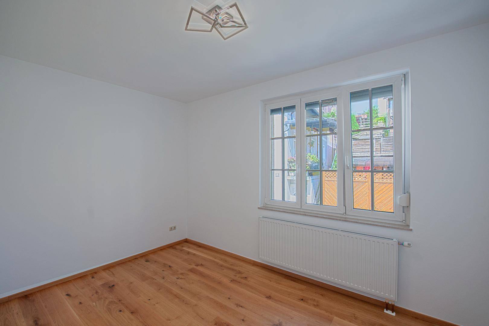 Leeres Zimmer mit Holzboden, weißen Wänden und einem Fenster, das viel Tageslicht hereinlässt.