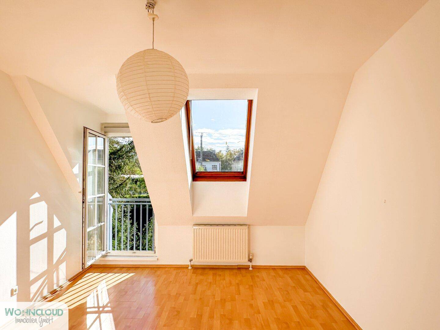 Helles Dachzimmer mit Parkettboden, Dachfenster und Zugang zu einem kleinen Balkon.