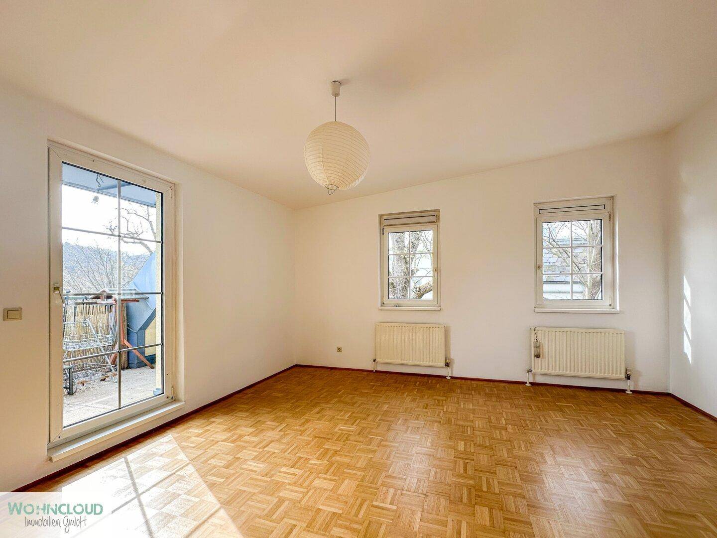 Lichtdurchfluteter Raum mit Parkettboden, zwei Fenstern und Balkonzugang.