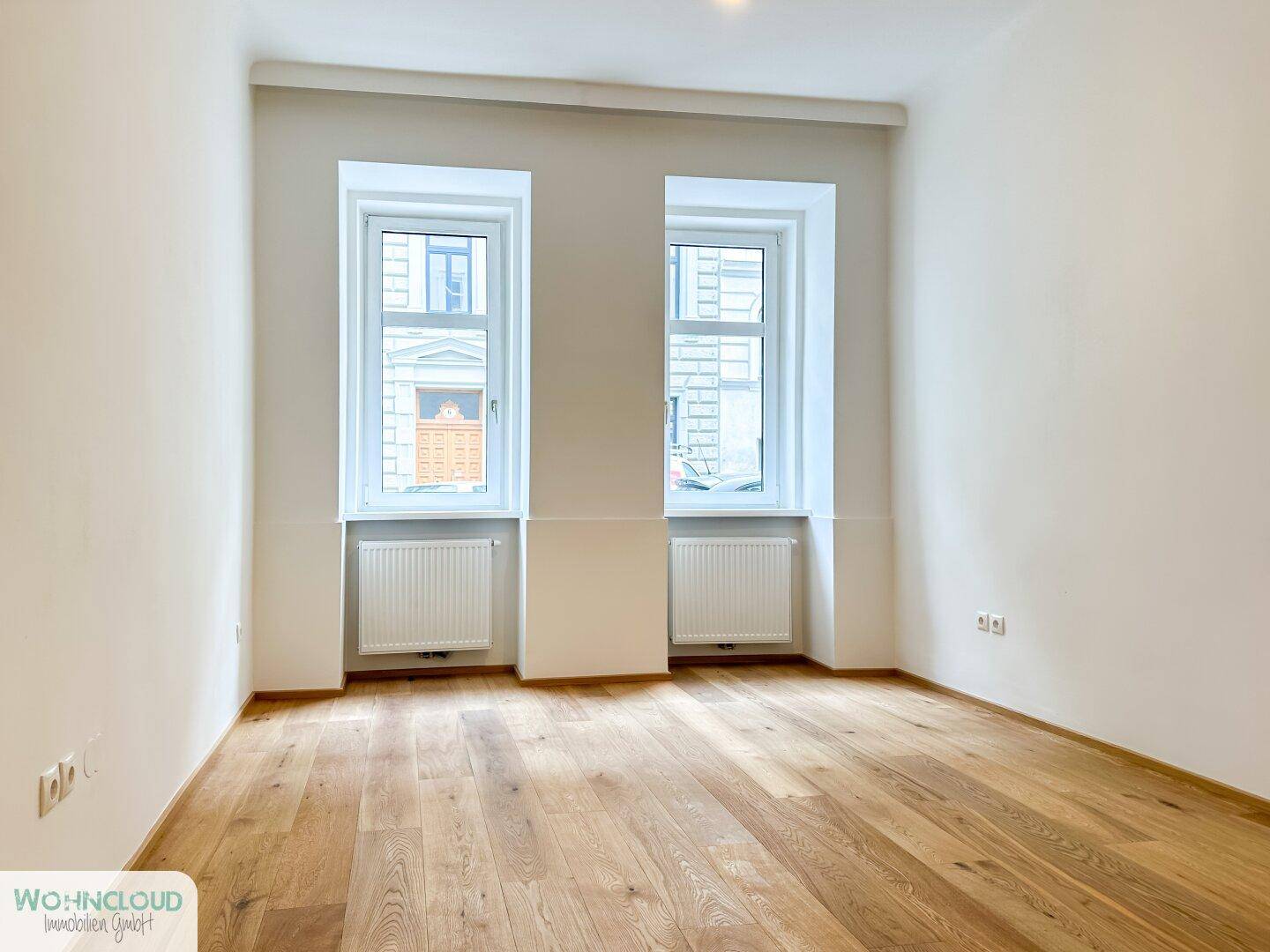 Leeres Zimmer mit Holzboden und zwei Fenstern, bereit zur individuellen Gestaltung und Nutzung.