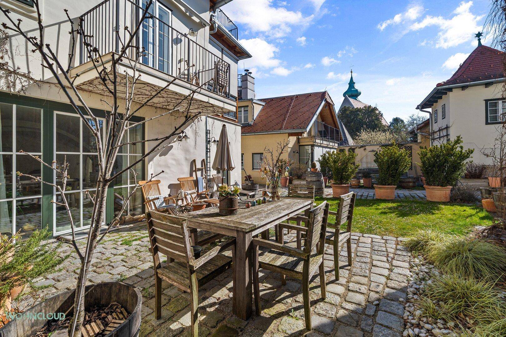 Sonniger Außenbereich mit gepflasterter Terrasse, Holztisch und Stühlen, umgeben von Grünflächen.