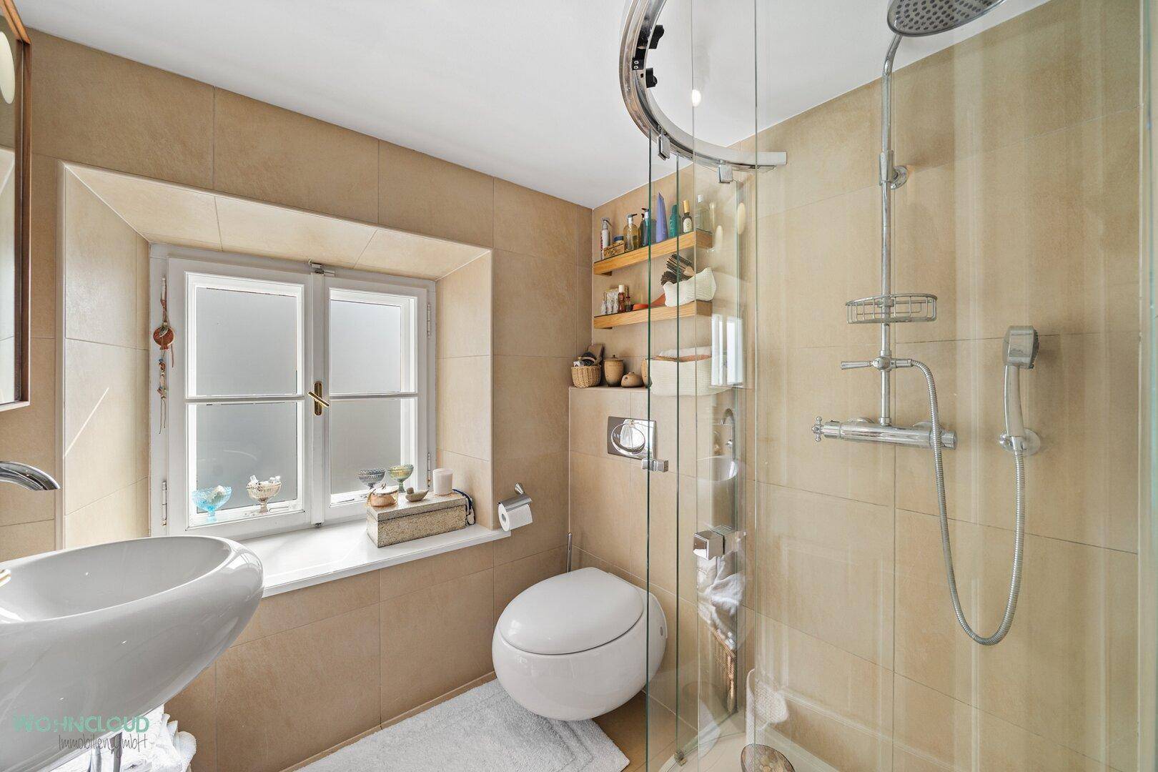 Modernes Badezimmer mit beigefarbenen Fliesen, Dusche, WC und Fenster für Tageslicht.