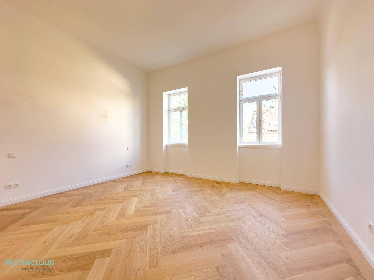 Helles Schlafzimmer mit klassischem Fischgrätparkett, weißen Wänden und zwei Fenstern für natürliche Belichtung.