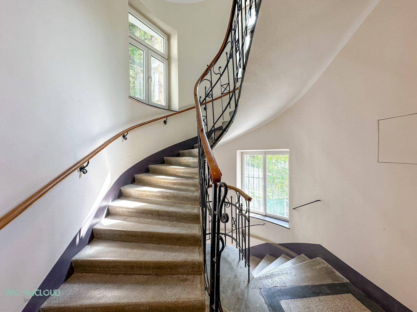 Historisches Treppenhaus mit geschwungenem Geländer, Steinstufen und großen Fenstern.