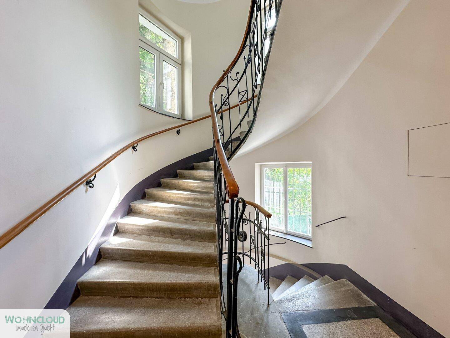 Elegantes Treppenhaus mit geschwungener Steintreppe, schmiedeeisernem Geländer und Fenstern.