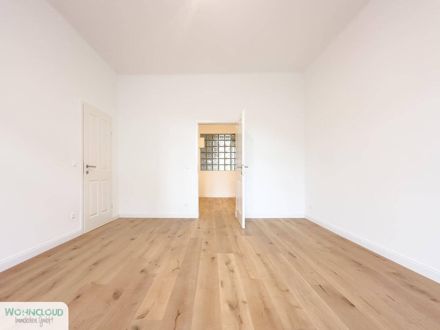Geräumiges, leeres Zimmer mit hellem Parkettboden und Durchgang zu einem weiteren Bereich.