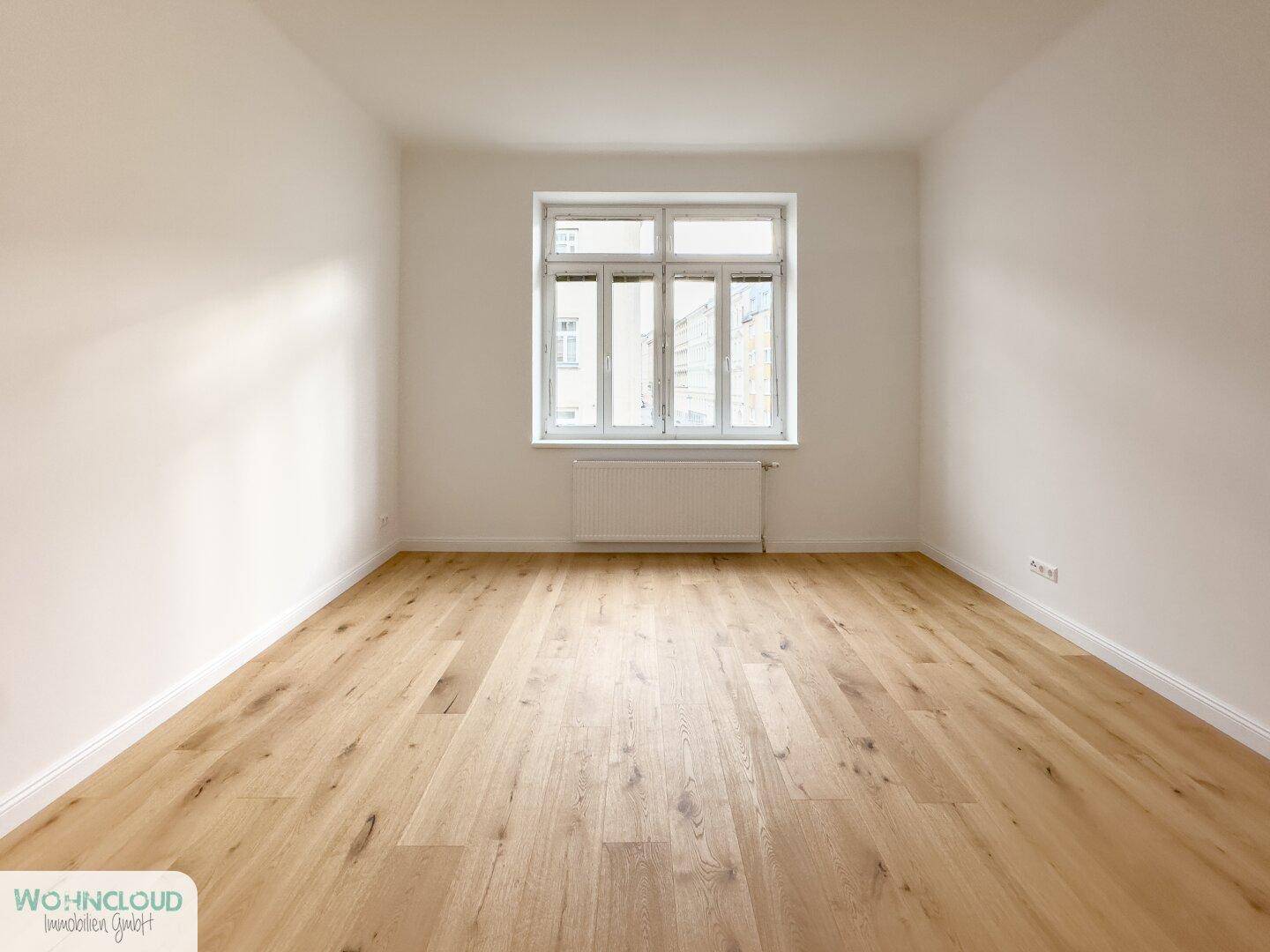 Helles, leeres Zimmer mit großem Fenster, Parkettboden und Heizkörper, ideal als Schlafzimmer.