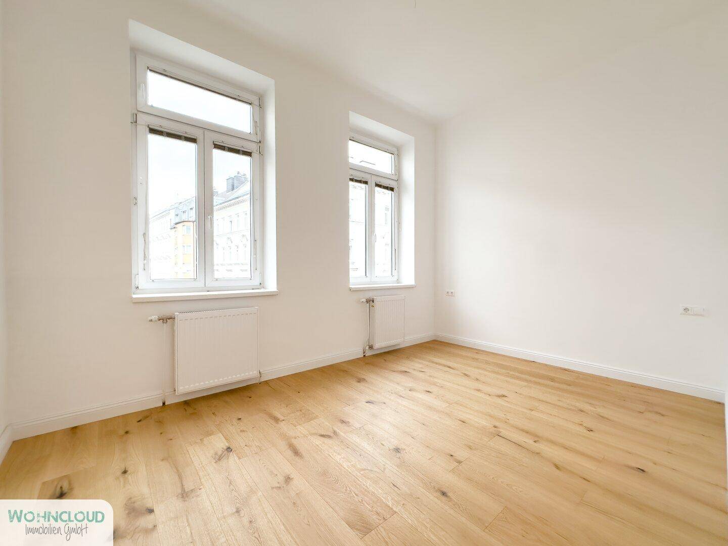 Lichtdurchflutetes Zimmer mit zwei großen Fenstern, Parkettboden und Heizkörpern.