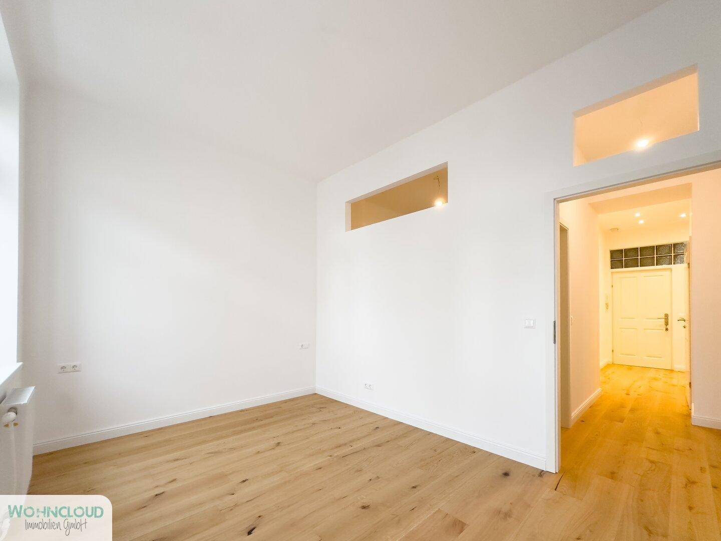 Leeres Zimmer mit Parkettboden und Durchgang zum Flur, bietet vielseitige Nutzungsmöglichkeiten.