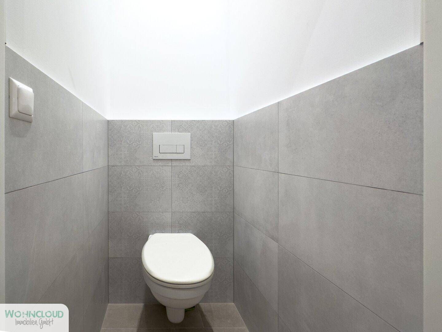 Separates WC mit wandhängender Toilette und modernen grauen Fliesen an Wänden und Boden.