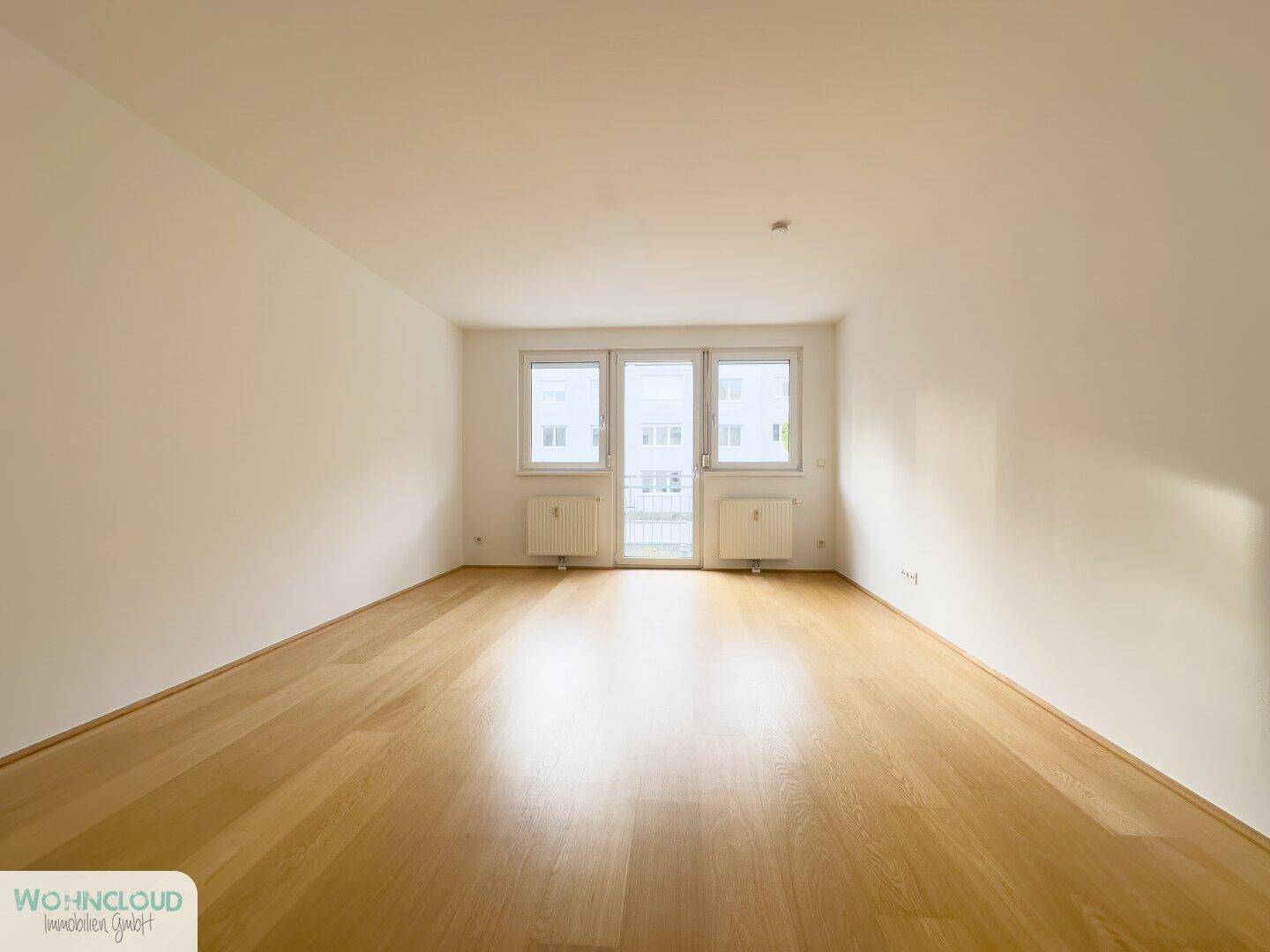 Geräumiger, leerer Wohnraum mit hellem Holzboden und Zugang zu einem französischen Balkon.