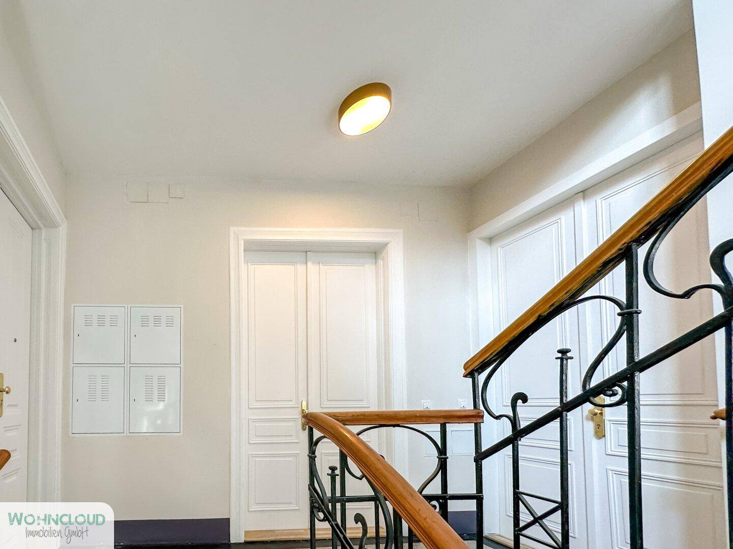 Heller Treppenaufgang in einem Altbau mit schmiedeeisernem Geländer und hohen Türen.