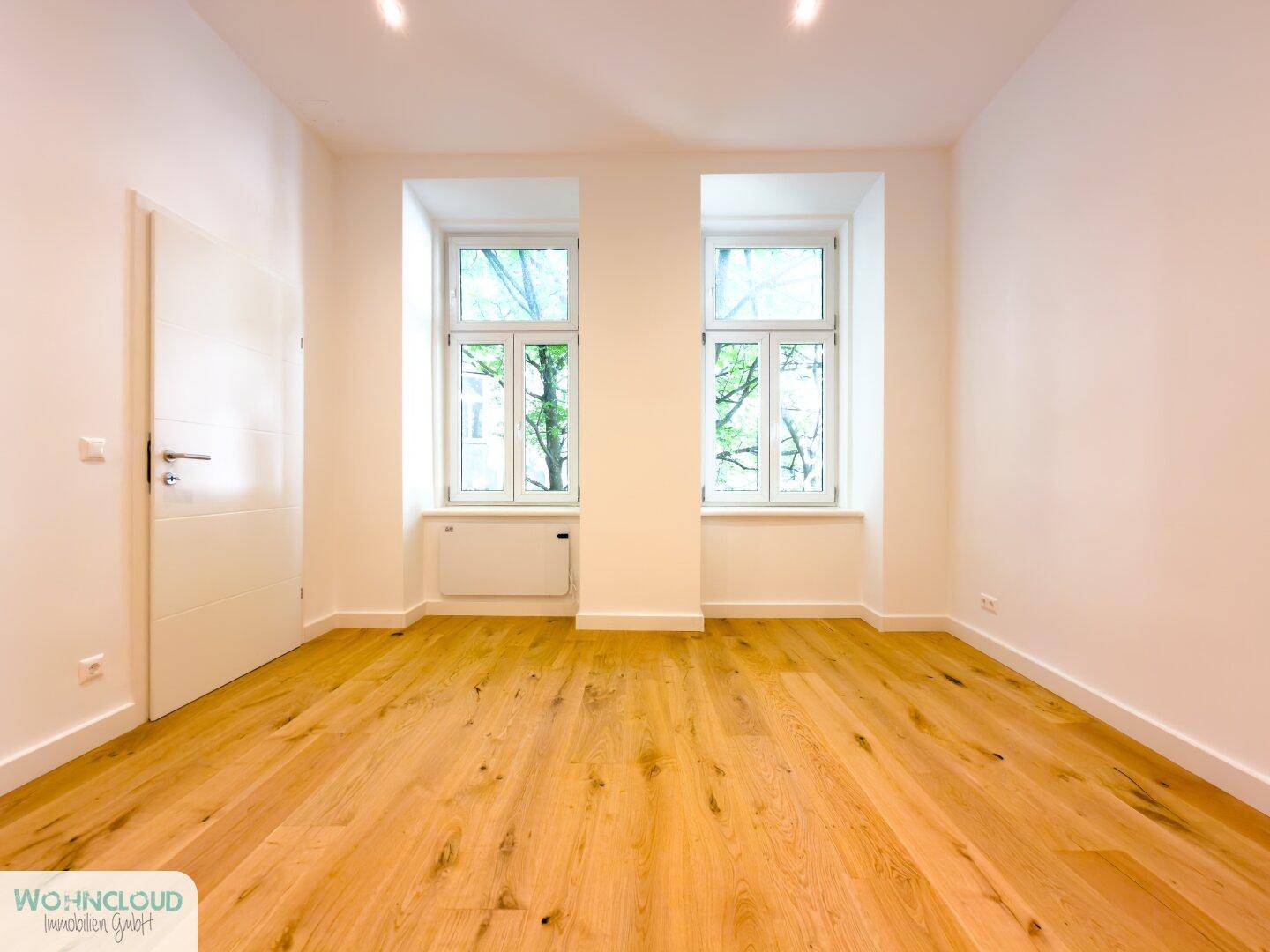 Lichtdurchflutetes, leeres Zimmer mit Holzboden und zwei Fenstern, ideal als Schlaf- oder Arbeitszimmer.