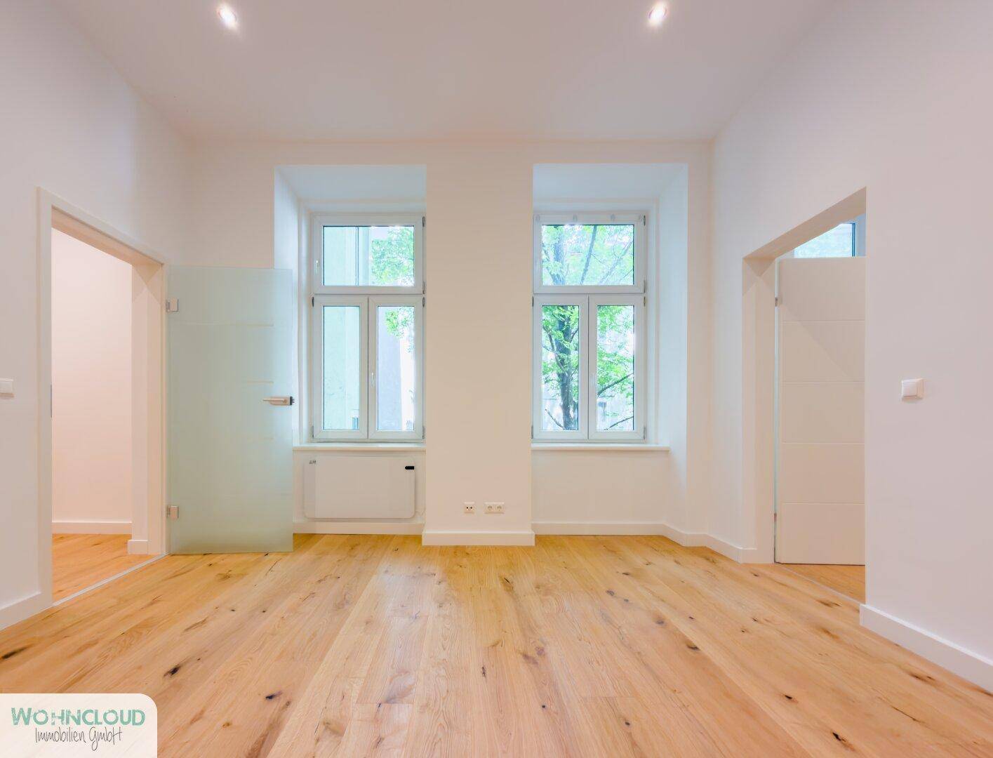 Geräumiges, leeres Zimmer mit hellem Holzfußboden und großen Fenstern für viel Tageslicht.