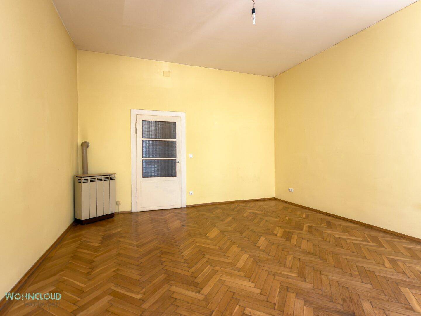 Geräumiges Zimmer mit gelben Wänden, Fischgrätparkett und einem Ofen, ideal für Wohnzwecke.