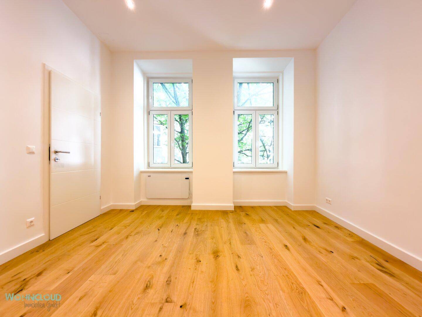 Helles, leeres Zimmer mit Holzboden und großen Fenstern, bereit zur individuellen Einrichtung.