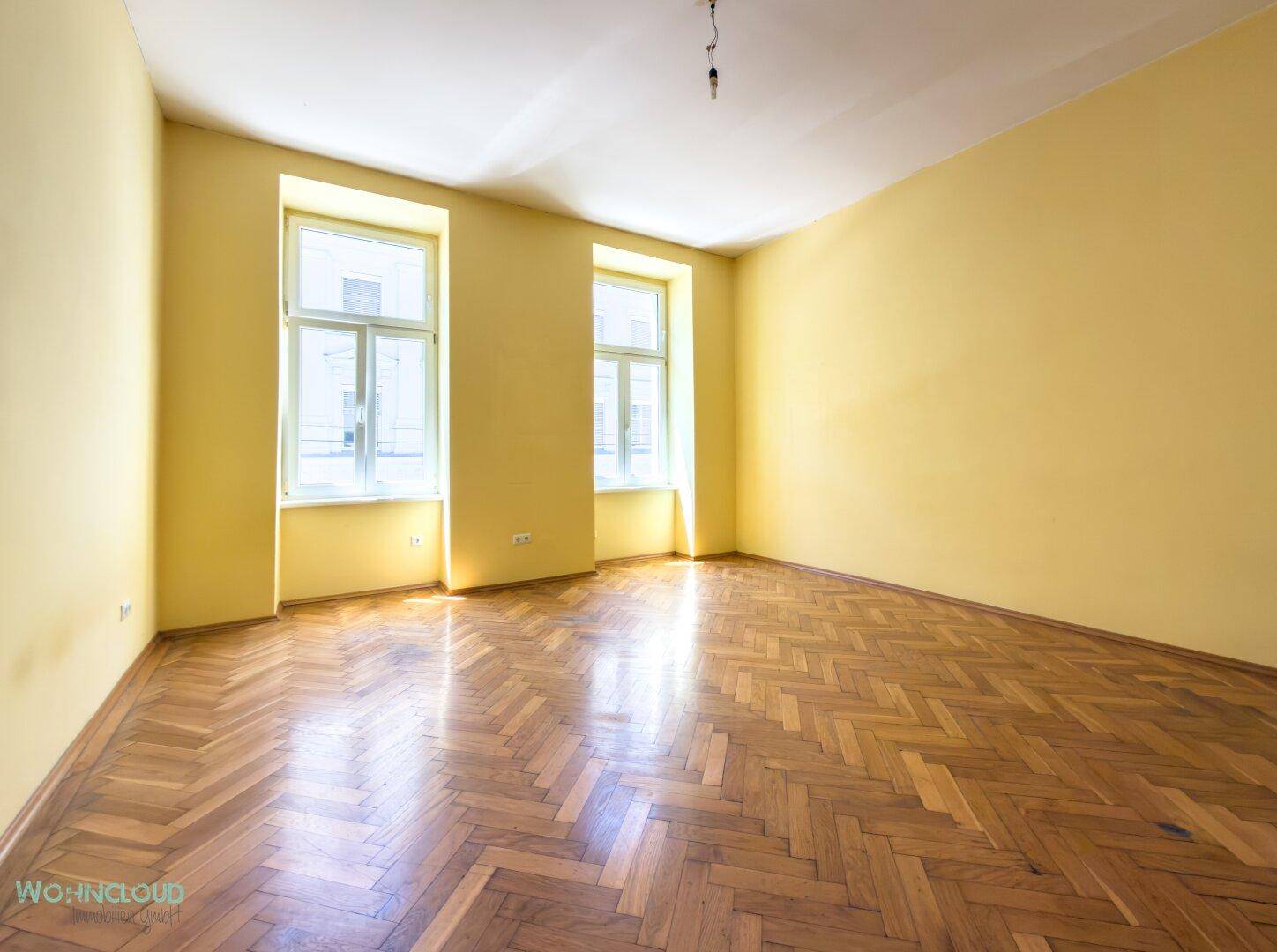 Geräumiges, leeres Zimmer mit gelben Wänden und glänzendem Fischgrätparkett.