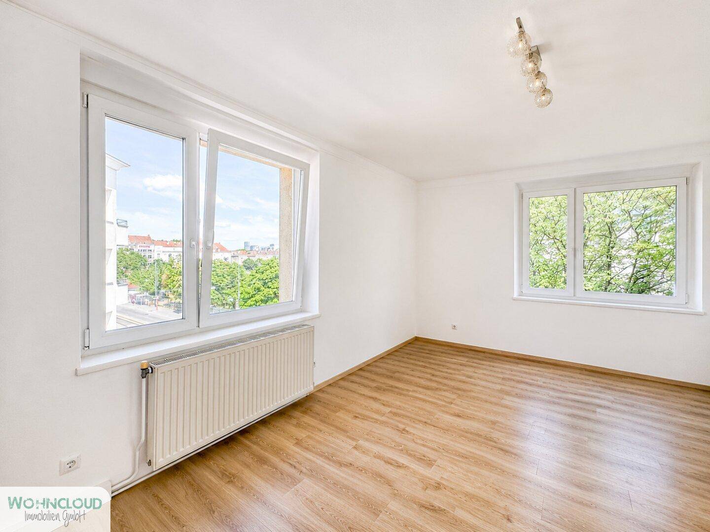 Helles Zimmer mit Laminatboden und zwei Fenstern, die einen Blick ins Grüne bieten.